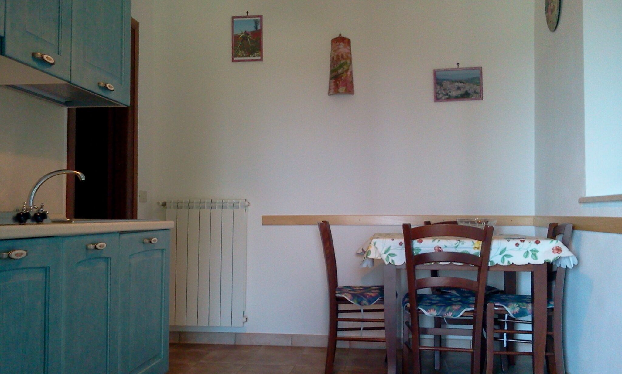 Une cuisine avec des meubles de couleur turquoise, un évier et un radiateur. On y trouve deux chaises recouvertes de nappes à motifs floraux et une chaise en bois avec un coussin à motifs.