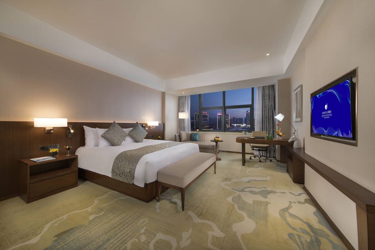 Een hotelkamer met een groot bed, een bureau, een televisie en uitzicht op de stad in de schemering.