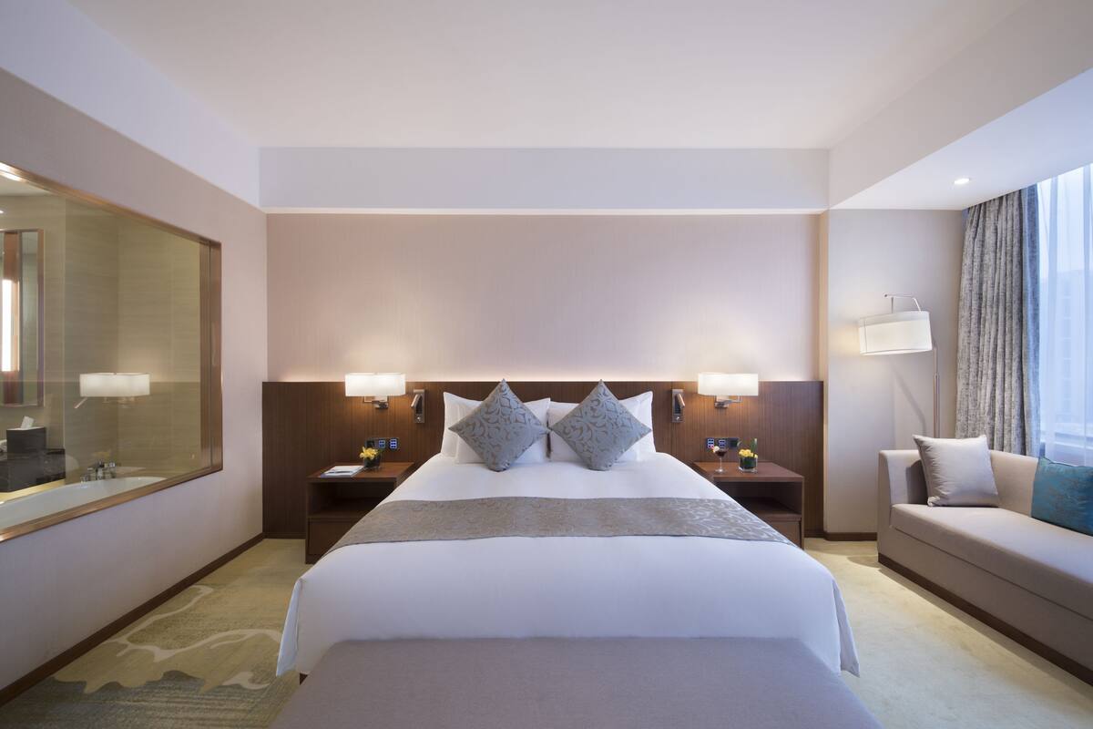 Hotelkamer met een groot bed, twee nachtkastjes, een bank en een grote spiegel, en een raam met gordijnen.