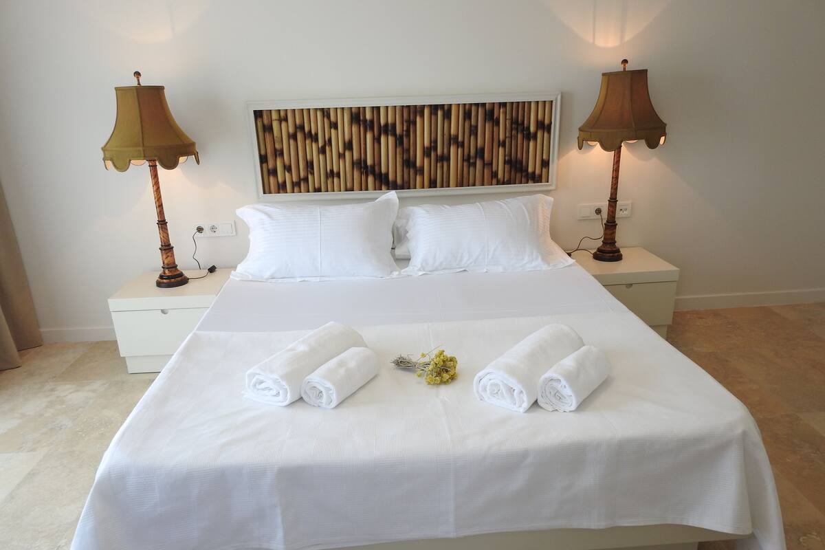 A neatly made bed with white linens, two bedside lamps, a wall-mounted artwork, and a small table with a lamp.