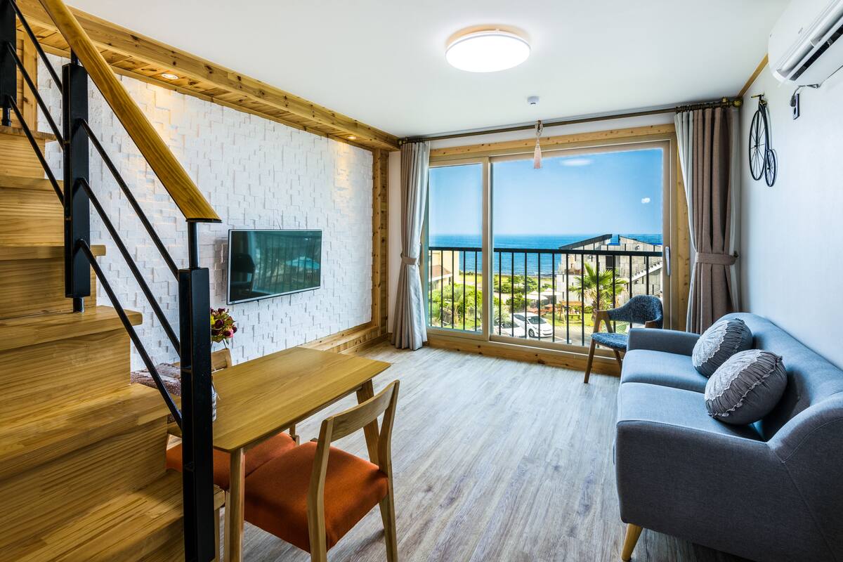 A modern living room with a wooden staircase, a dining area, and a balcony with a view of the sea.