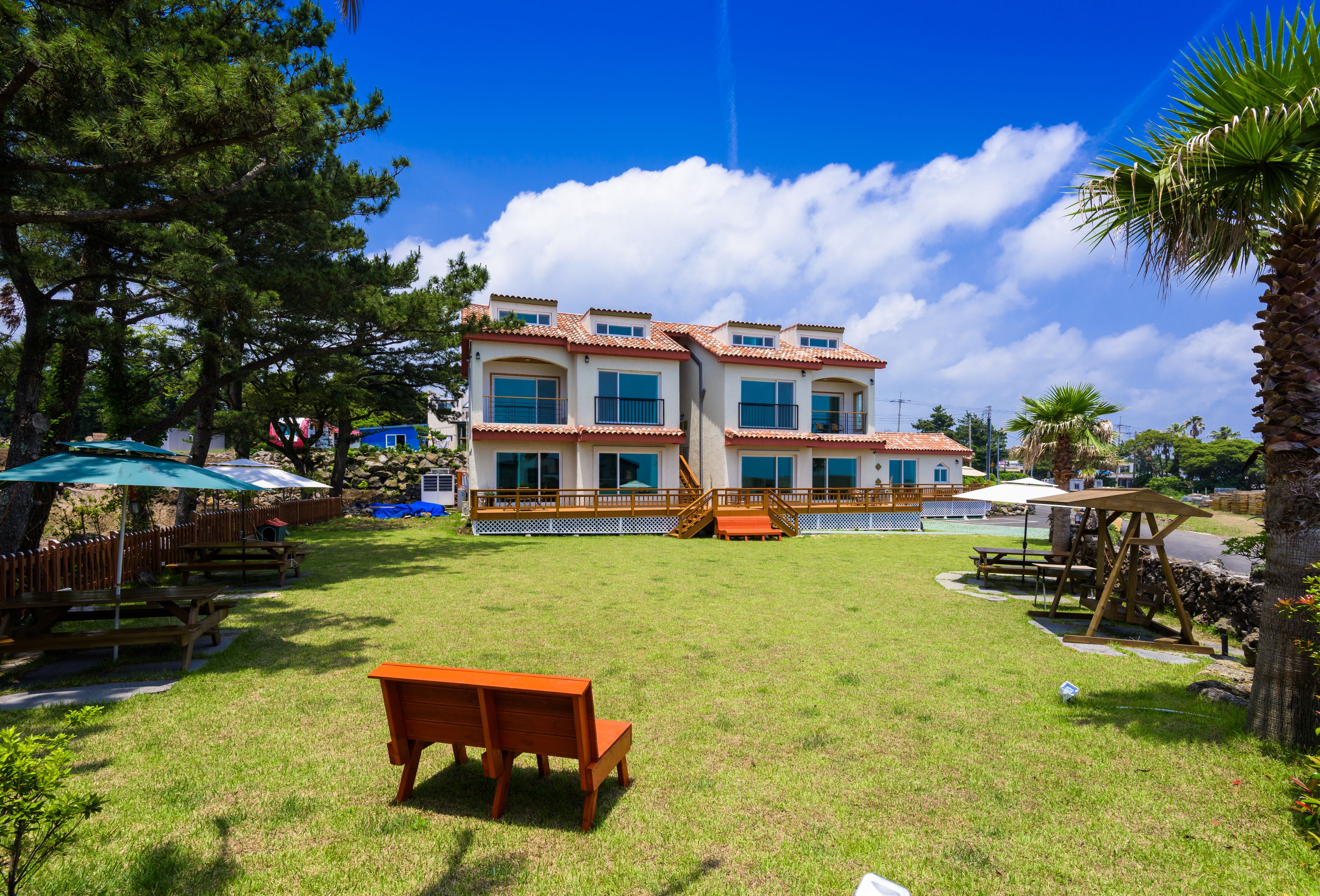 A two-story building with a tiled roof, a covered patio area, and a grassy lawn with a wooden bench.