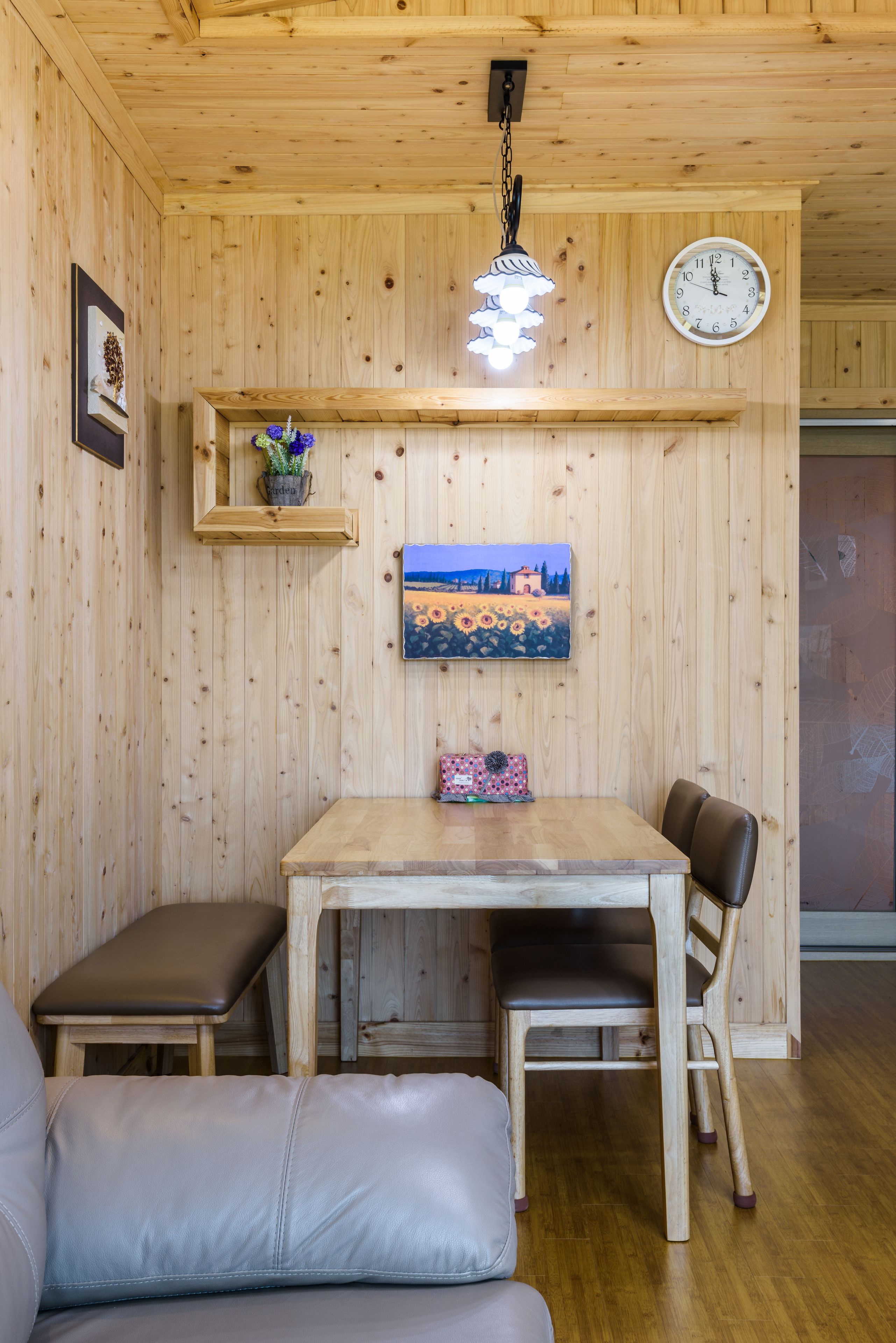 Een kamer met houten lambrisering, een houten eettafel, twee stoelen, een bankje, een klok en een wandplank met een vaas en een schilderij.