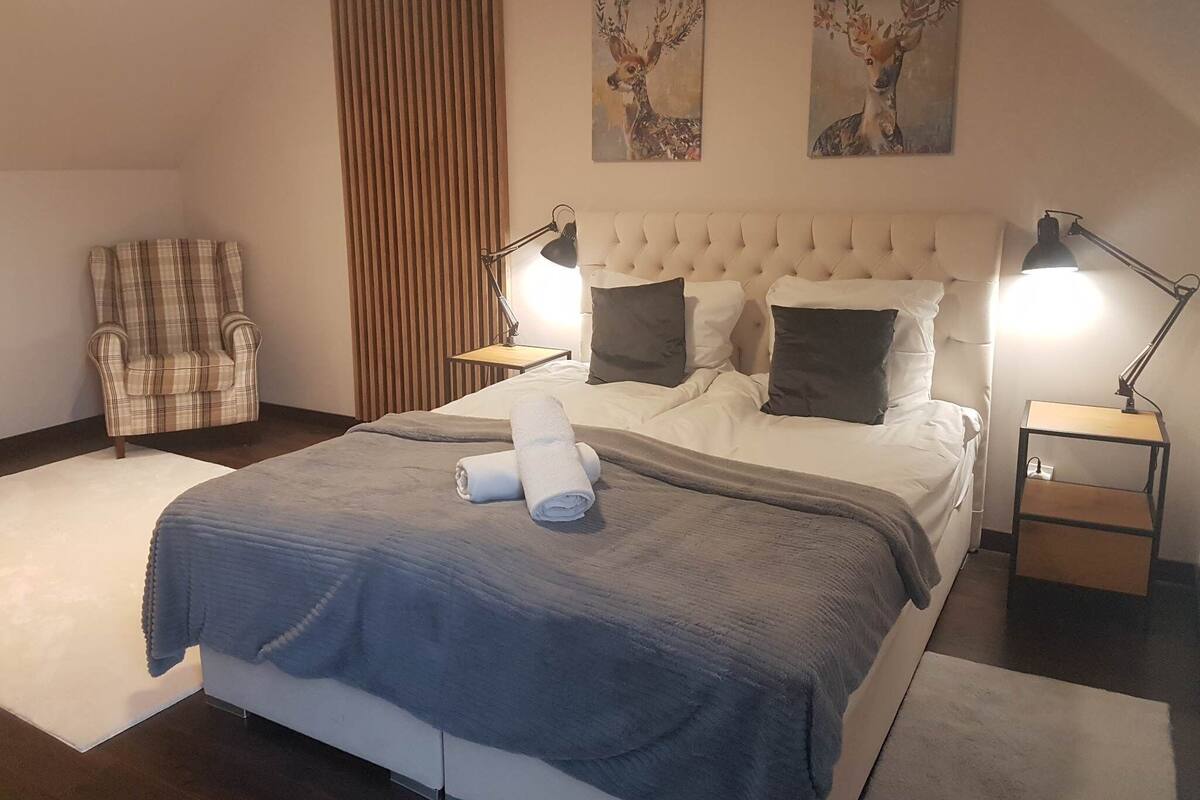Ein ordentlich bezogenes Bett mit blauer Decke, zwei Nachttischlampen und ein karierter Sessel.