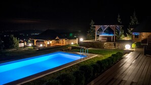 Ein Nachtblick auf einen Pool mit einer Holzterrasse, einem Gazebo und einem Steinhaus.