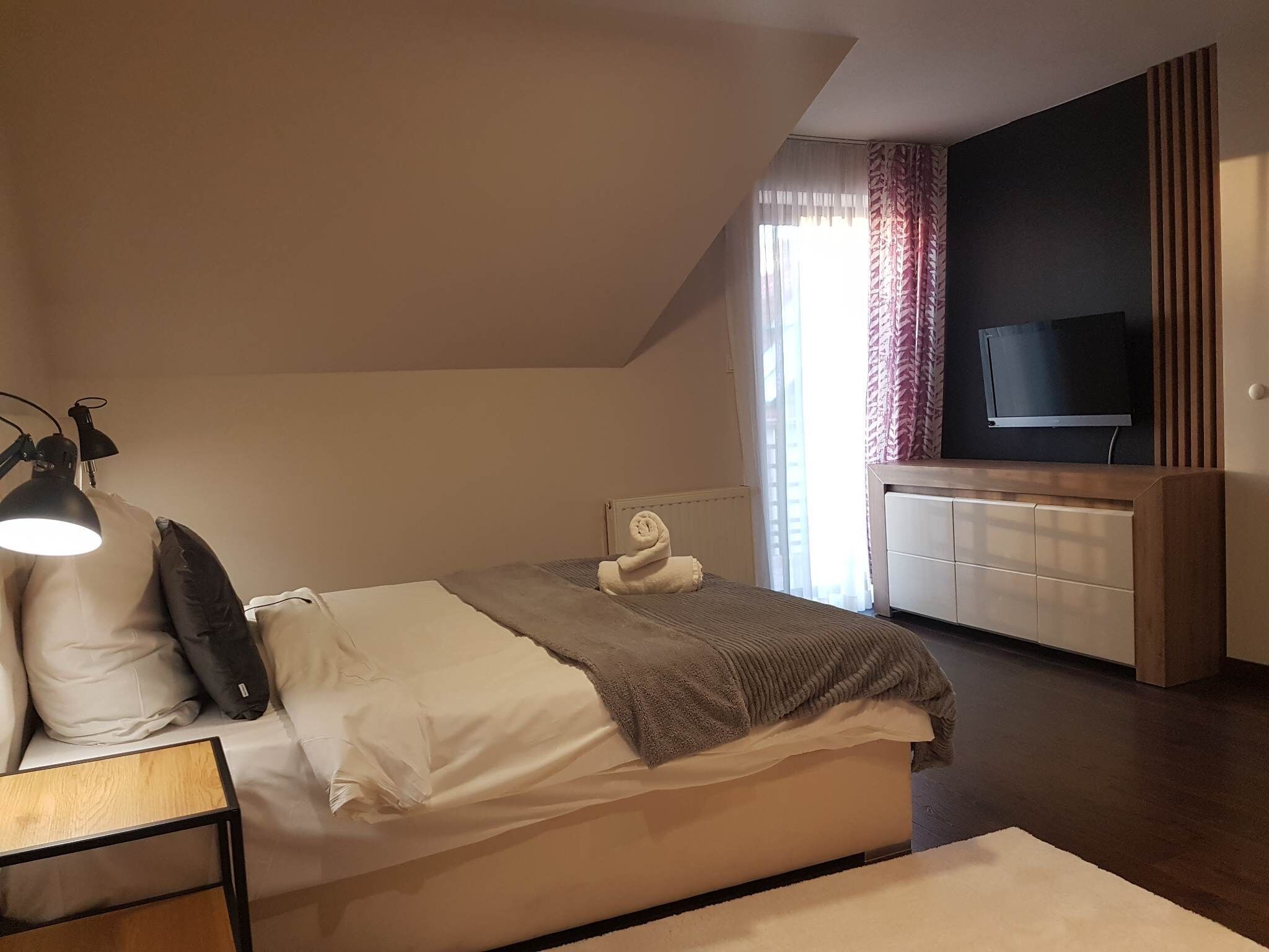 Een slaapkamer met een bed, bedlampje, een televisie op een houten standaard en een raam met gordijnen.