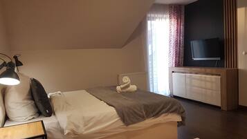 Een slaapkamer met een bed, bedlampje, een televisie op een houten standaard en een raam met gordijnen.