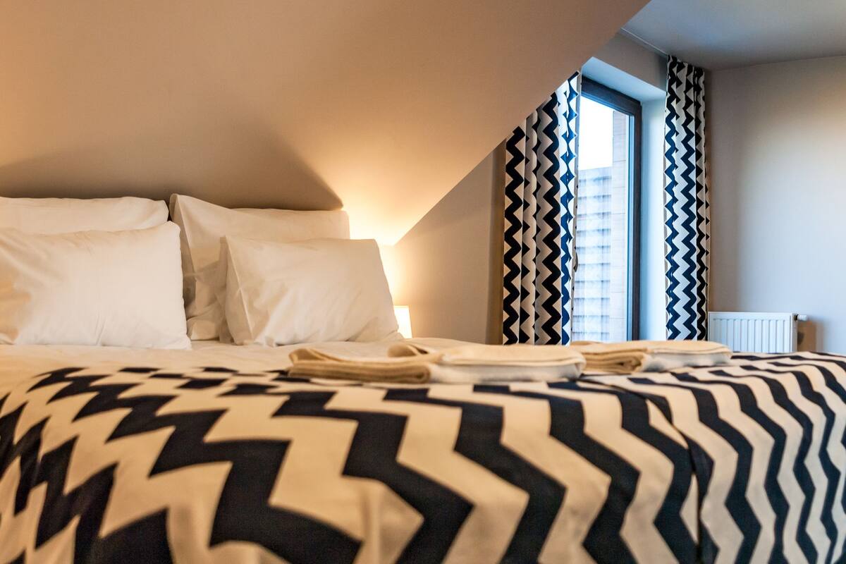 Ein Hotelzimmer mit Bett, Nachttisch, Lampe, Kleiderschrank und einem Fenster mit Vorhängen.