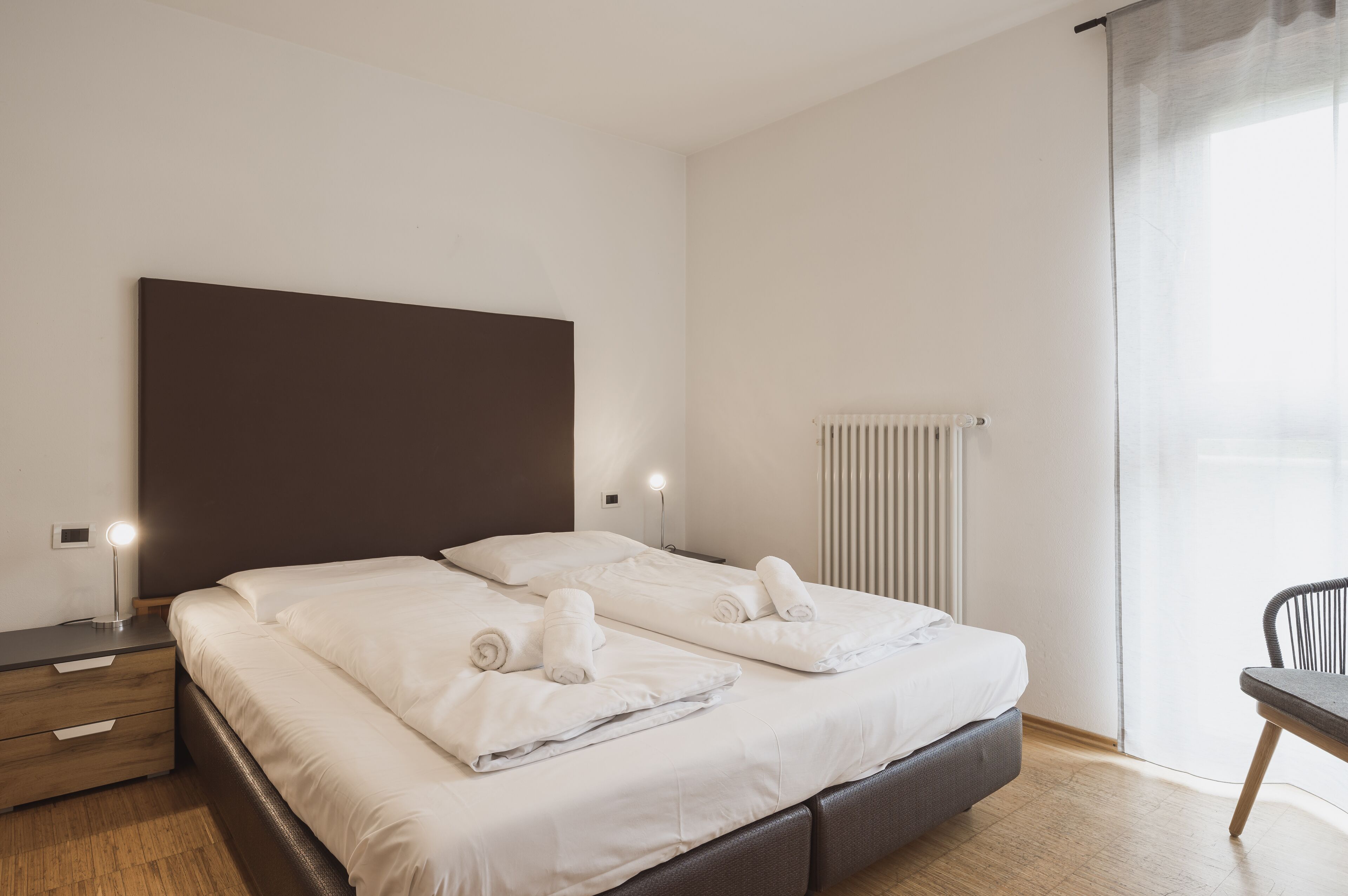 A double bed with white linens and rolled towels, a bedside table with a lamp, a radiator, and a chair.