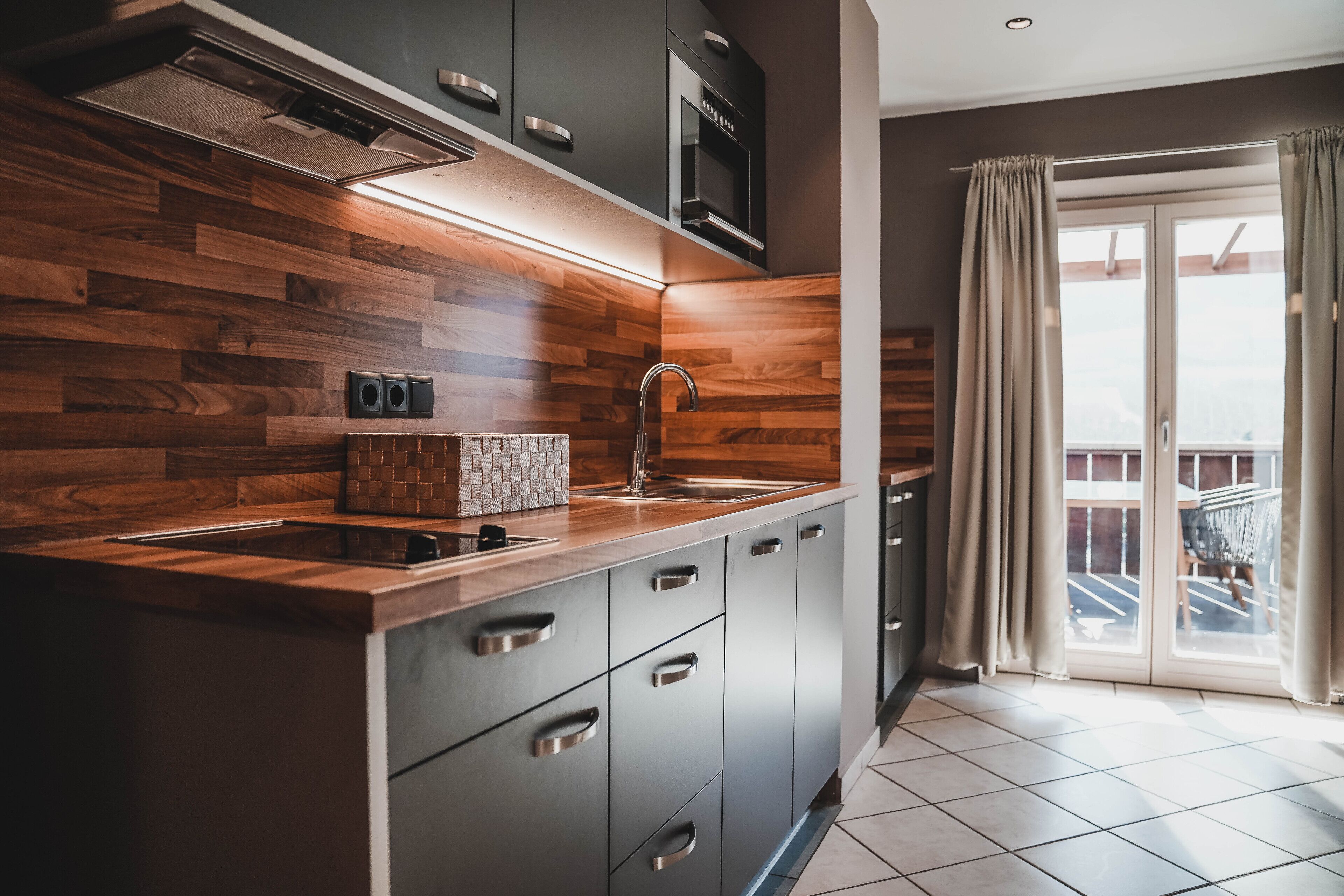 A modern kitchen with wooden cabinetry, a built-in microwave, and a large window with curtains.