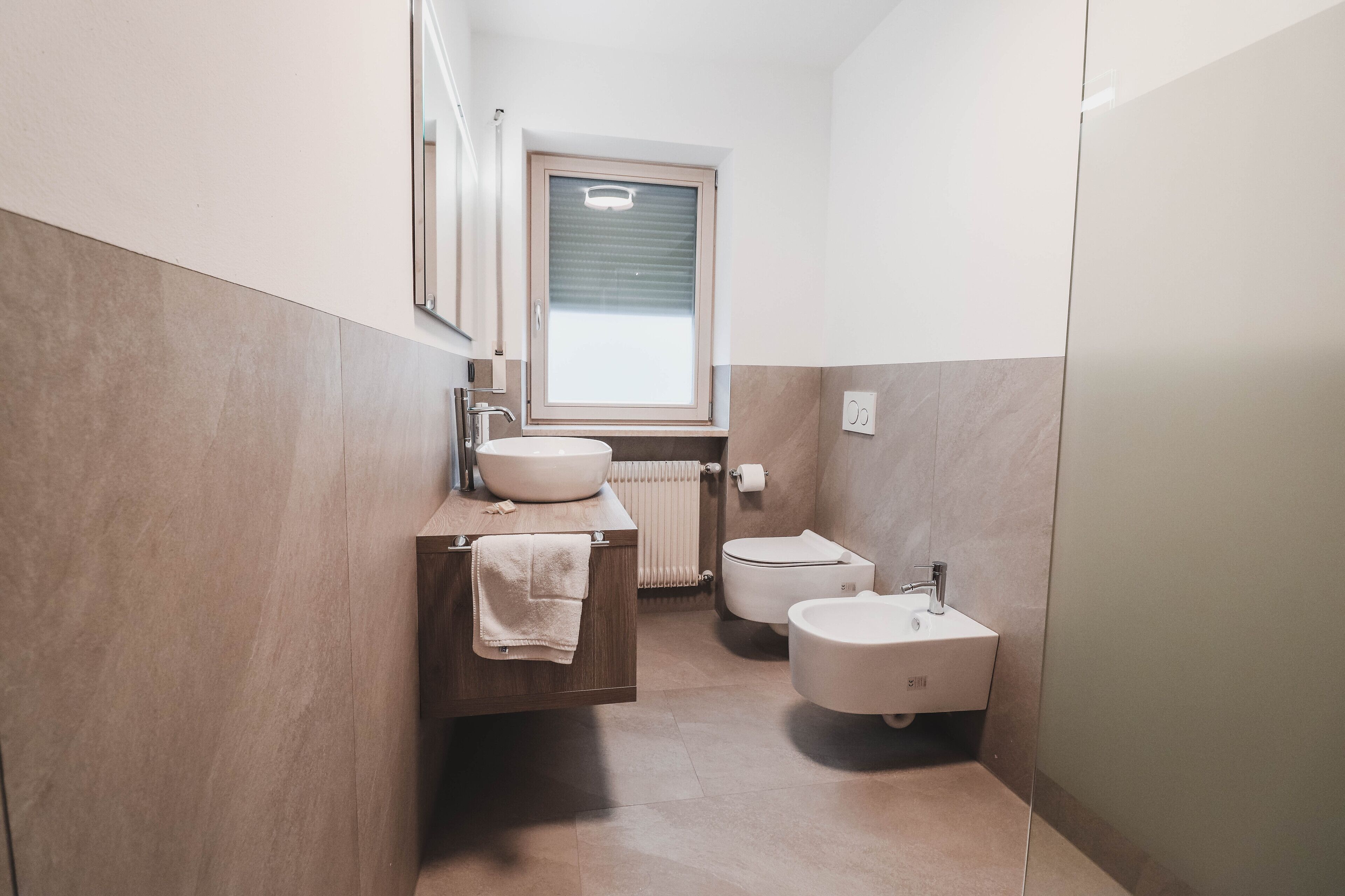 A compact bathroom with a wooden vanity, a large mirror, a window, and two sinks.