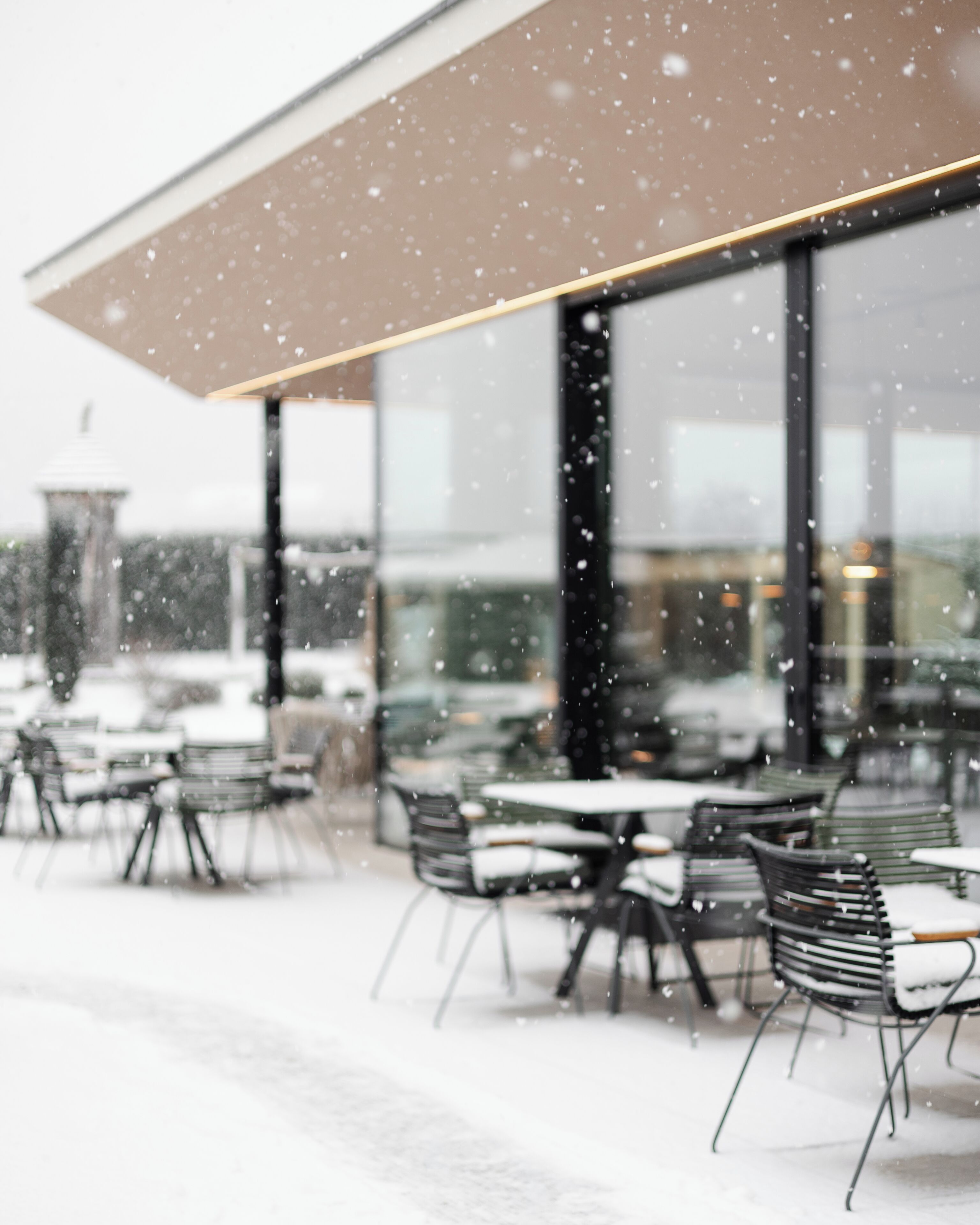 Un café al aire libre con mesas y sillas, grandes ventanales y suelo cubierto de nieve.