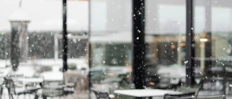 An outdoor cafe with tables and chairs, large windows, and a snow-covered ground.