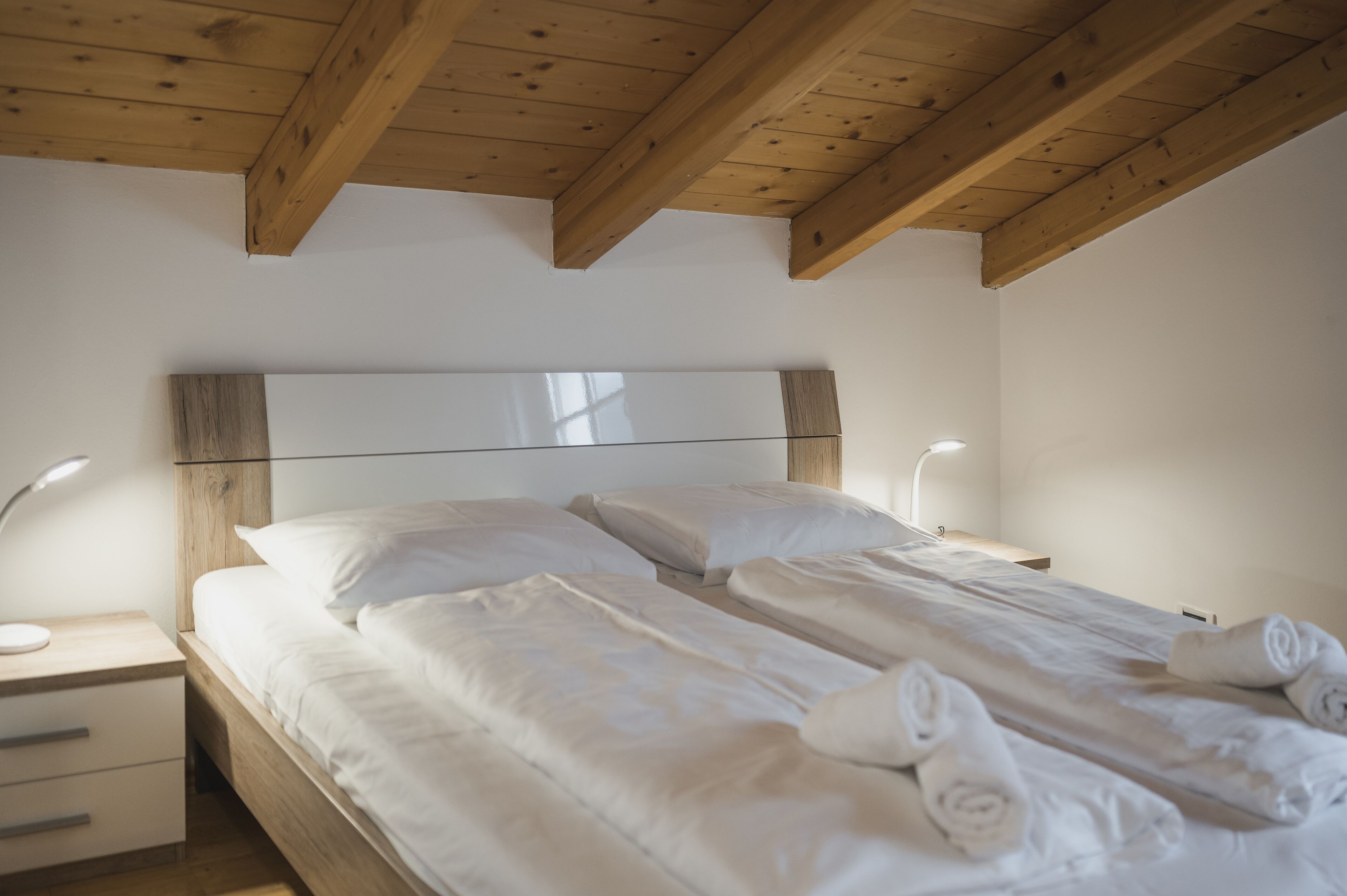 Un letto matrimoniale con lenzuola bianche e asciugamani arrotolati, una testiera in legno, comodini con lampade e un soffitto in legno.