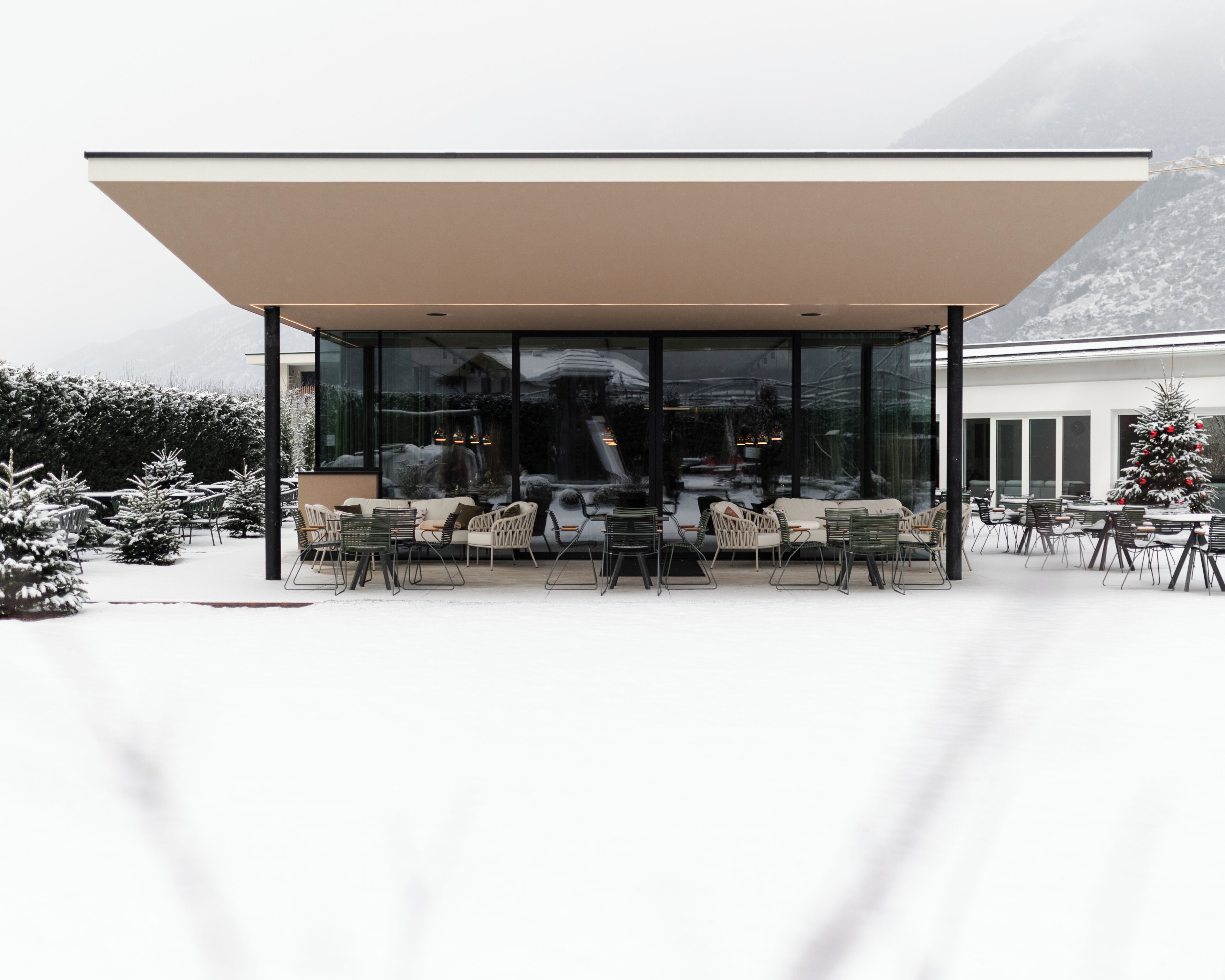 A modern building with a glass facade and a covered outdoor seating area with chairs and tables.