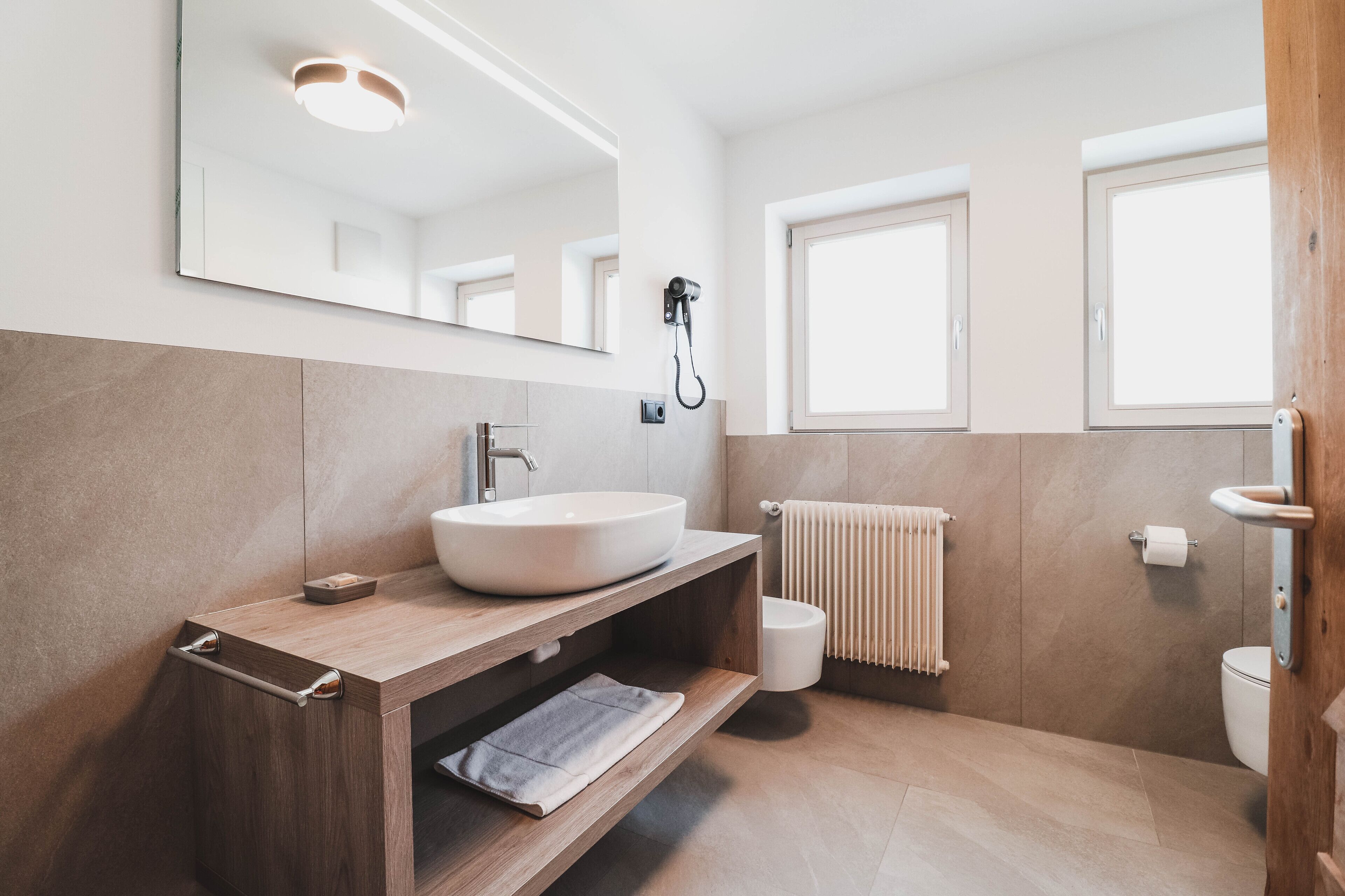 Une salle de bain moderne avec un grand miroir, un meuble-lavabo en bois, un lavabo blanc, un porte-serviettes, des toilettes et une fenêtre.