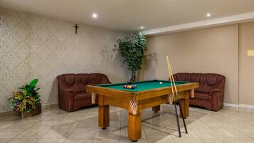 A pool table in a room with two leather chairs, a potted plant, and a cross on the wall.