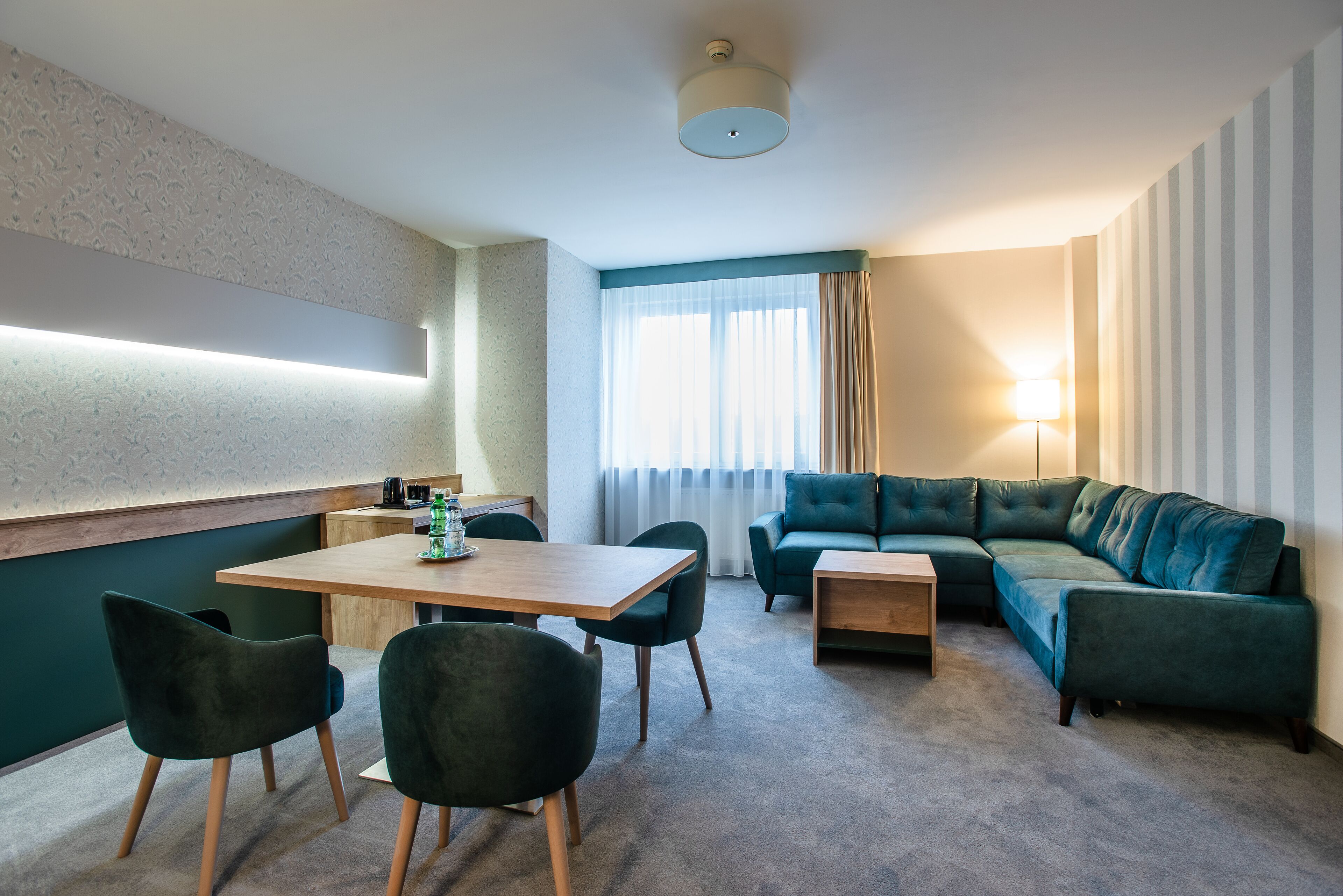 A modern hotel room with a wooden dining table, two green chairs, a green sofa, and a small side table.
