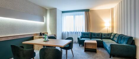 A modern hotel room with a wooden dining table, two green chairs, a green sofa, and a small side table.