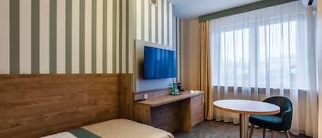 A hotel room with a bed, a desk with a TV, a chair, and a window with curtains.