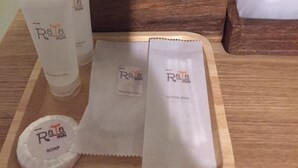Una bandeja de madera con artículos de tocador, incluyendo una botella de jabón, un tubo de loción y hisopos, colocada sobre una mesa con una caja de madera y una bolsa de plástico al fondo.