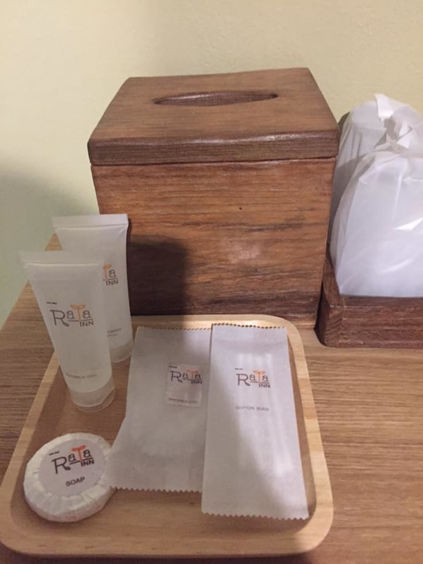 Una bandeja de madera con artículos de tocador, incluyendo una botella de jabón, un tubo de loción y hisopos, colocada sobre una mesa con una caja de madera y una bolsa de plástico al fondo.