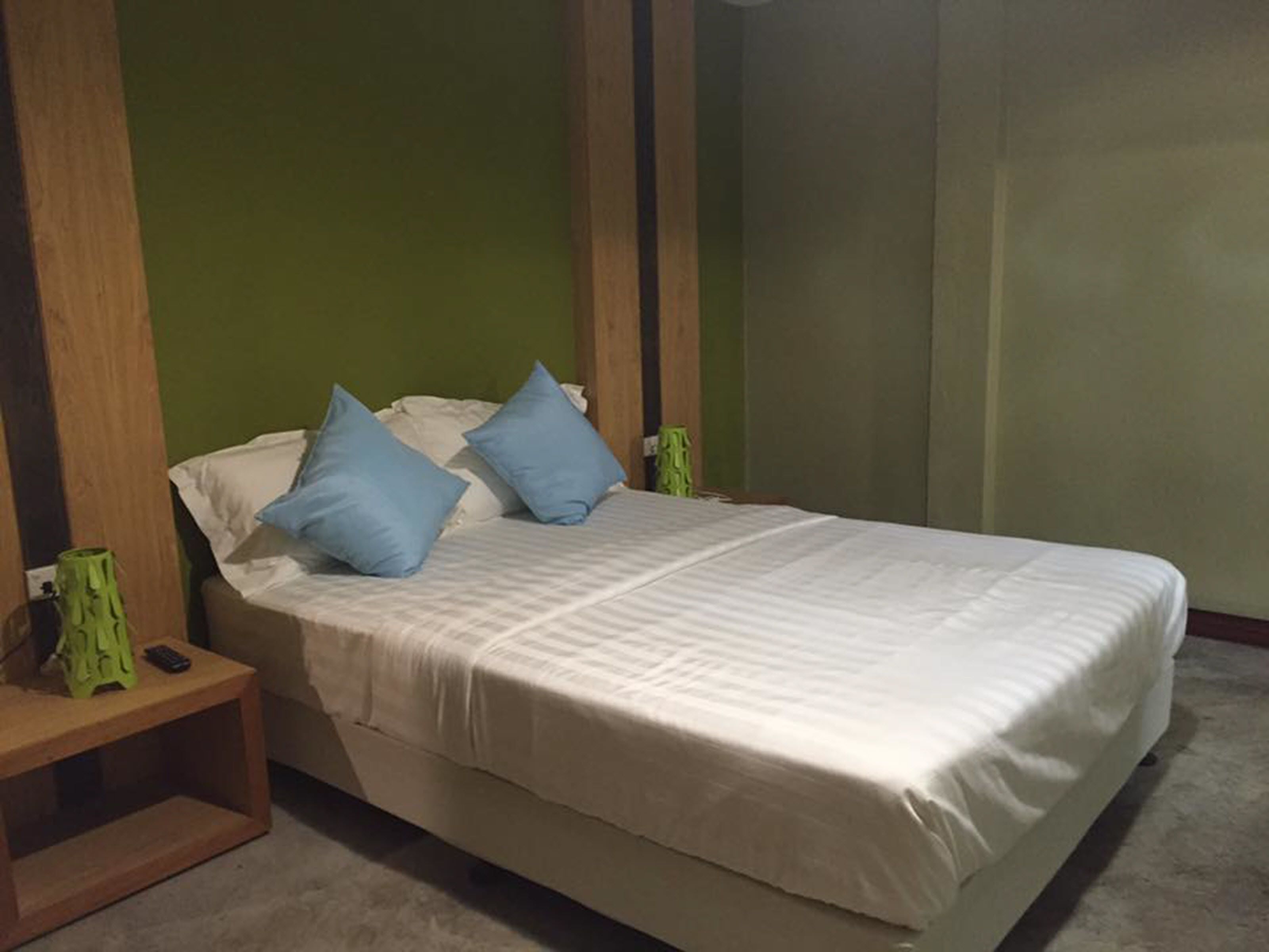 A single bed with a white mattress and blue pillows, a wooden nightstand with a green vase, and a green wall.