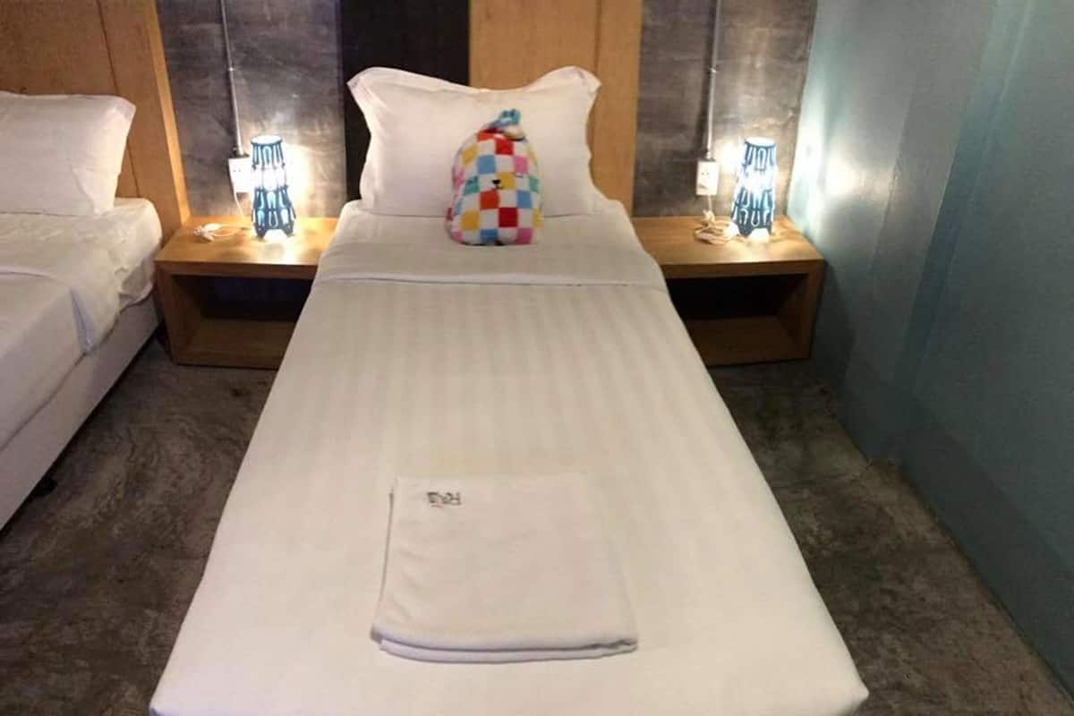 A hotel room with a single bed, two bedside tables with lamps, a colorful pillow, and a folded towel on the bed.