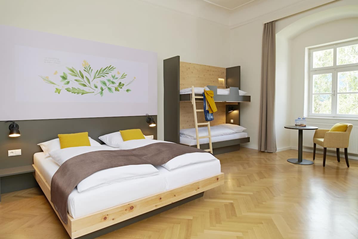 A modern hotel room with a wooden bed, a bunk bed, a small table with chairs, and a large window.