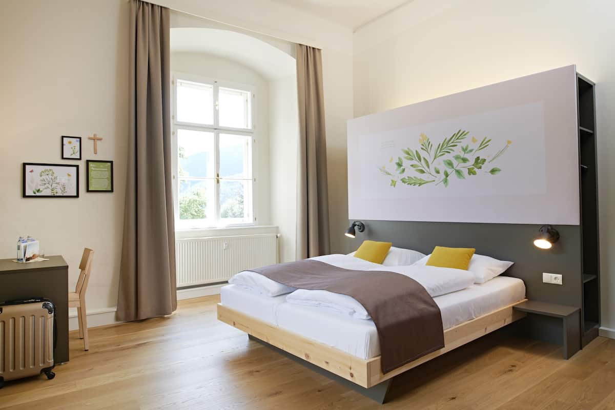 A modern bedroom with a wooden bed, a large window with curtains, a radiator, and a wall-mounted light fixture.
