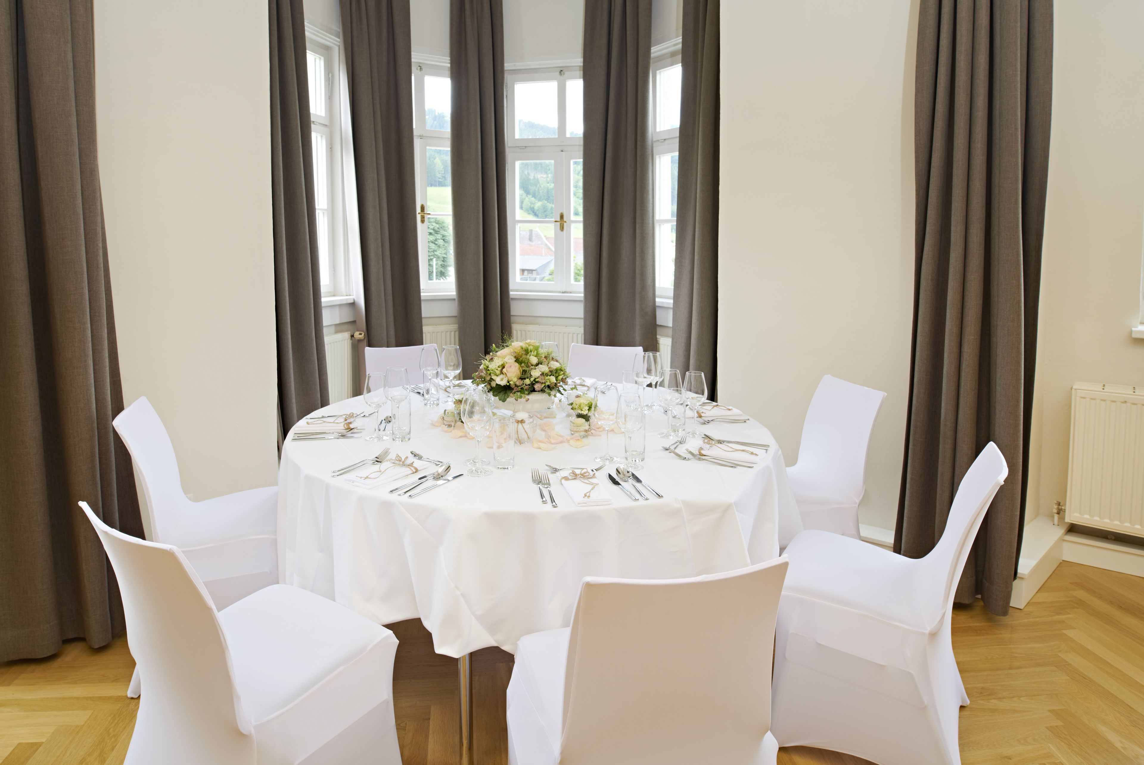 Une table ronde dressée pour six personnes, avec des chaises blanches, un arrangement floral au centre et une vue sur un bâtiment à travers de grandes fenêtres.