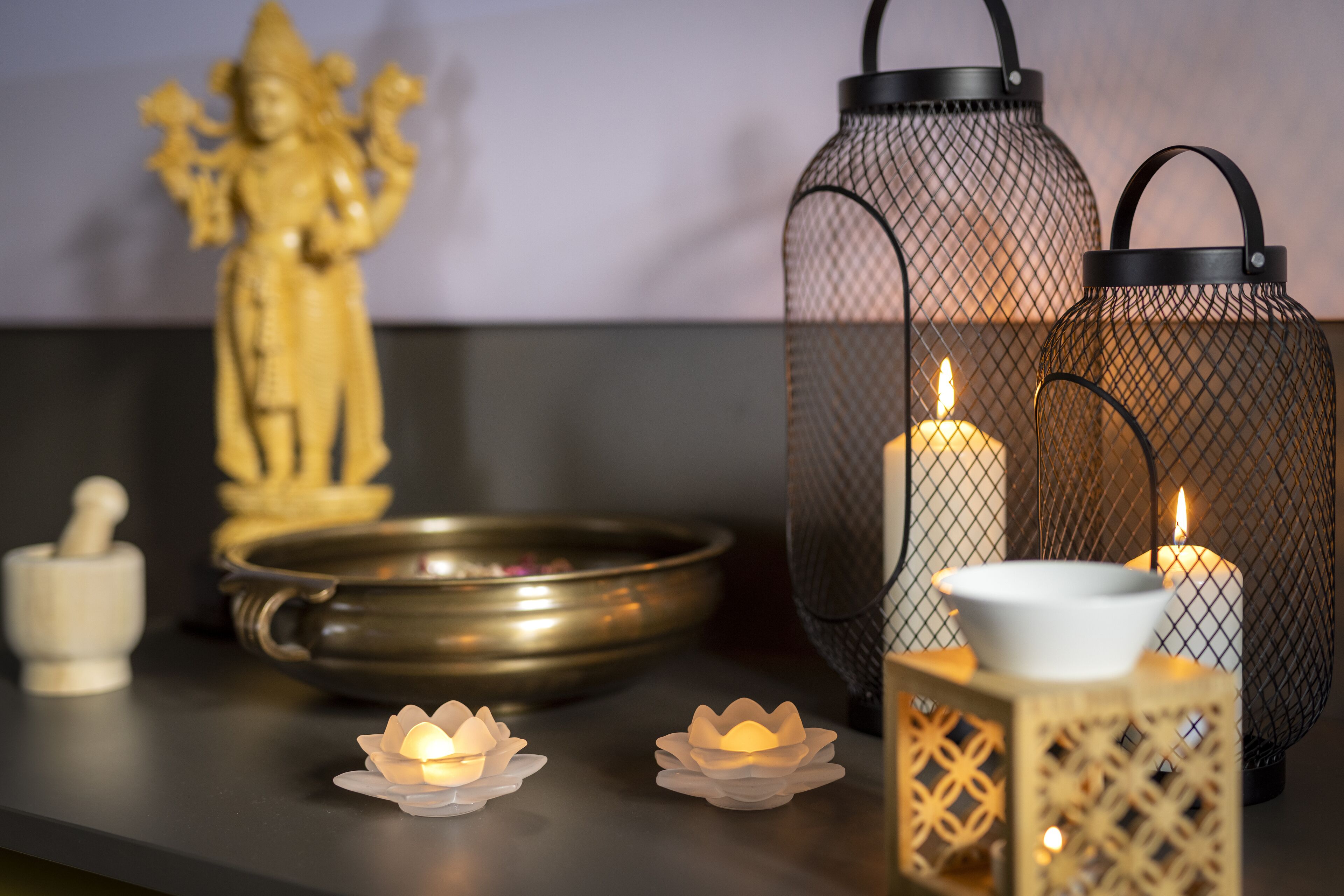 A table with a statue, a brass bowl, two lit candles, and two wire lanterns.