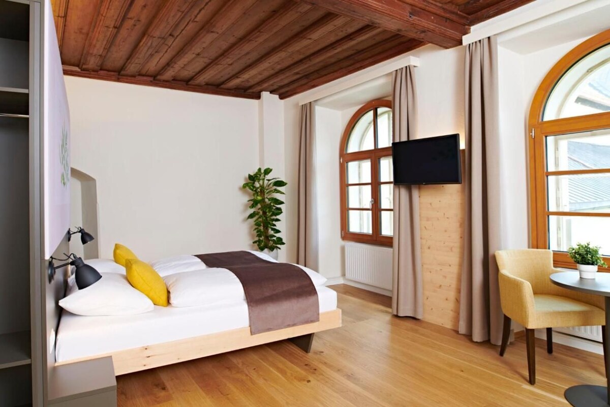 A modern bedroom with a wooden ceiling, a bed with a brown blanket, a TV on the wall, and a window with curtains.
