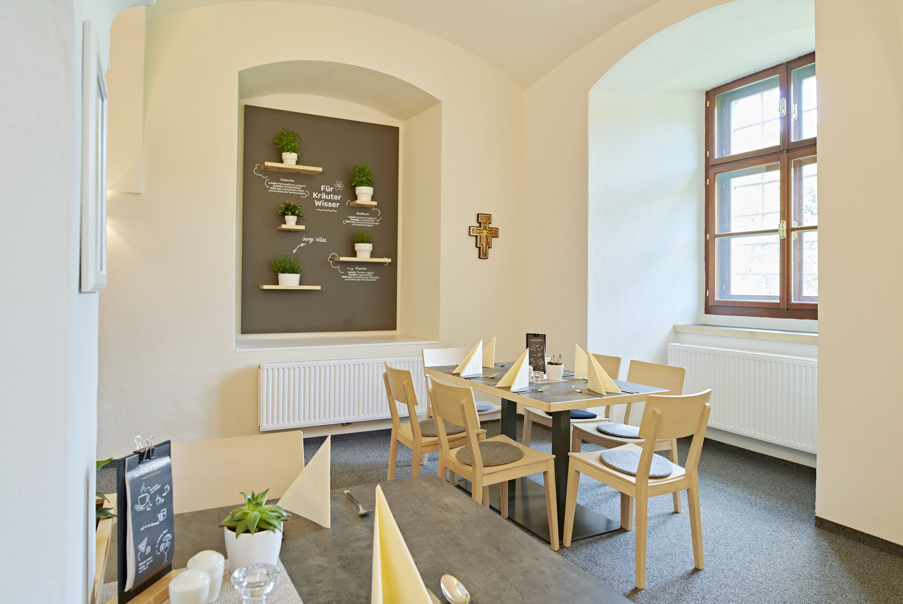 A dining area with a round table, chairs, a blackboard with text, a cross on the wall, and a window.