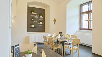 A dining area with a round table, chairs, a blackboard with text, a cross on the wall, and a window.
