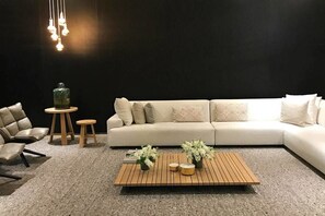 Uma sala de estar moderna com um sofá bege, uma mesa de centro de madeira com flores, uma poltrona de couro e uma pequena mesa de madeira com um vaso verde.