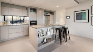 A modern kitchen with a central island, bar stools, and a view of the city through large windows. - Melbourne Docklands Concavo Seaview Apartment (Docklands)