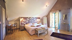 Een slaapkamer met een schuine kap, een bed met een bloemrijk hoofdeinde, nachtkastjes, een stoel, een bureau, een spiegel en gordijnen.