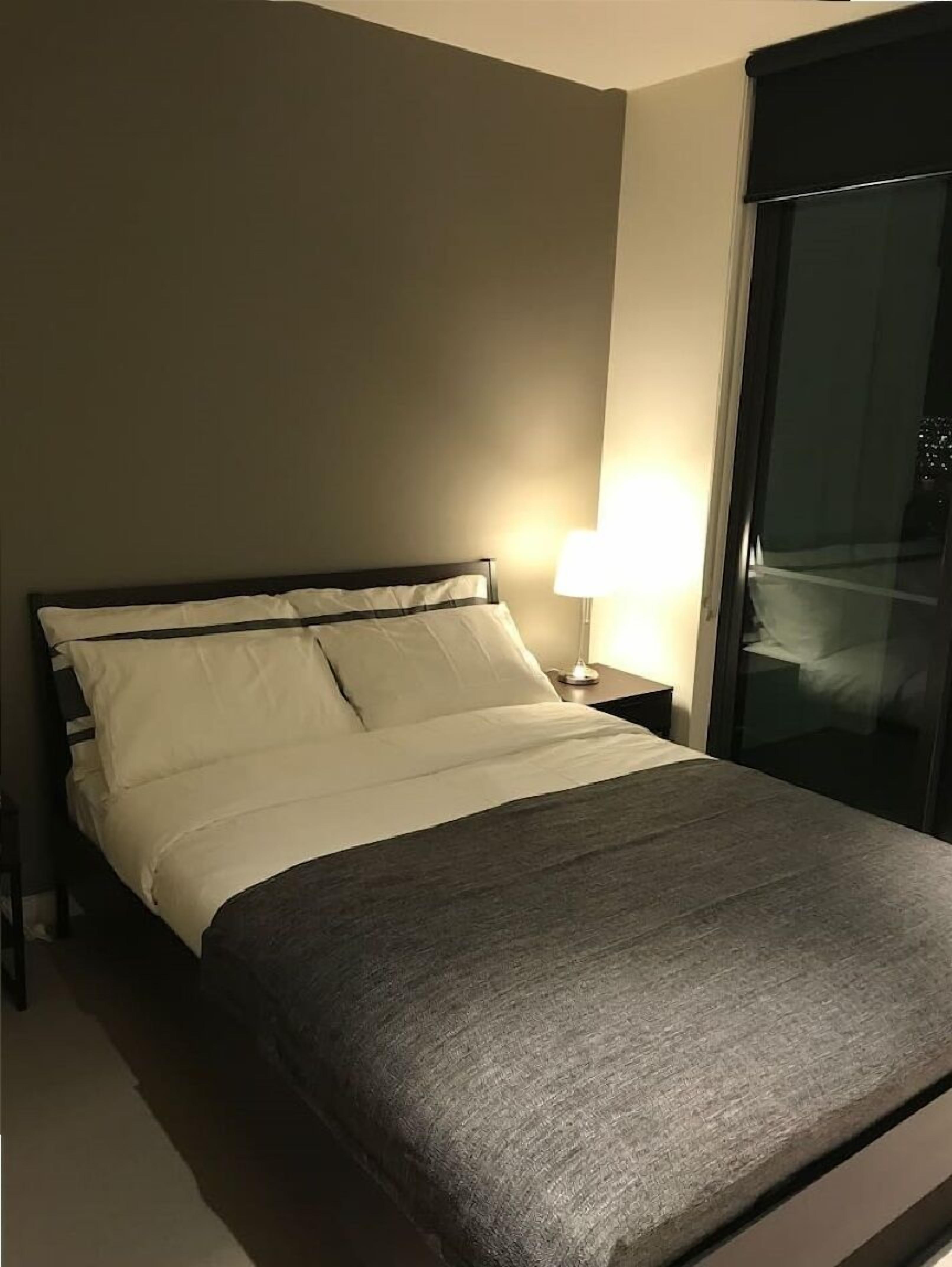 Cama bem arrumada com lençóis brancos e manta cinza, abajur e vista para uma varanda através de uma porta de vidro.