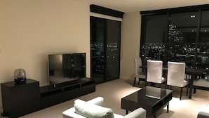 A modern living room with a flat-screen TV, a coffee table, and a sofa. There is a dining area with chairs and a table. The room has large windows with a city view. - Melbourne Docklands Convesso Seaview Apartment (Docklands)