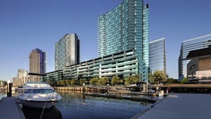 A modern building with a glass facade, a marina with boats, and other high-rise buildings in the background. - Melbourne Docklands Convesso Seaview Apartment (Docklands)