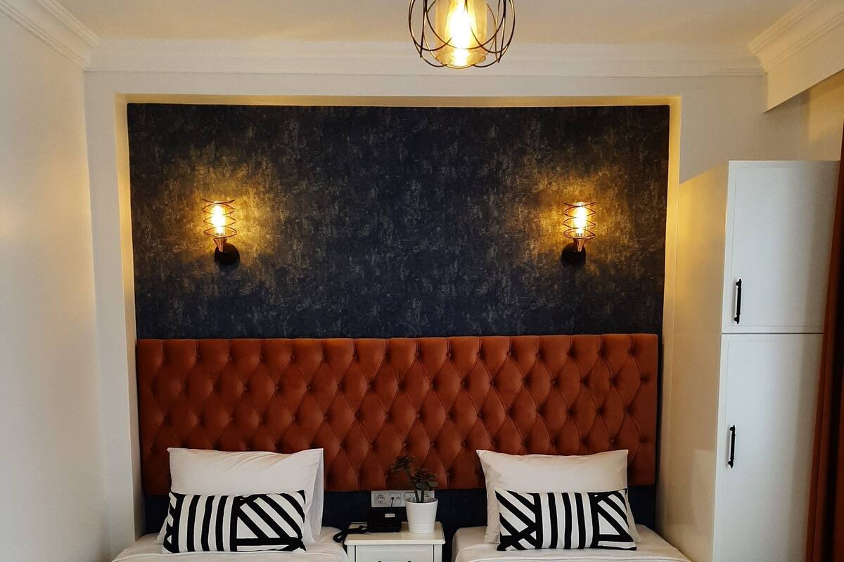 Une chambre d’hôtel avec deux lits, une tête de lit capitonnée, une petite table de chevet avec une radio et une armoire.