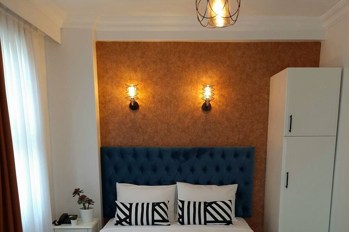 Une chambre d’hôtel avec un grand lit, deux lampes fixées au mur, une armoire blanche et une table de chevet avec un téléphone et une plante.