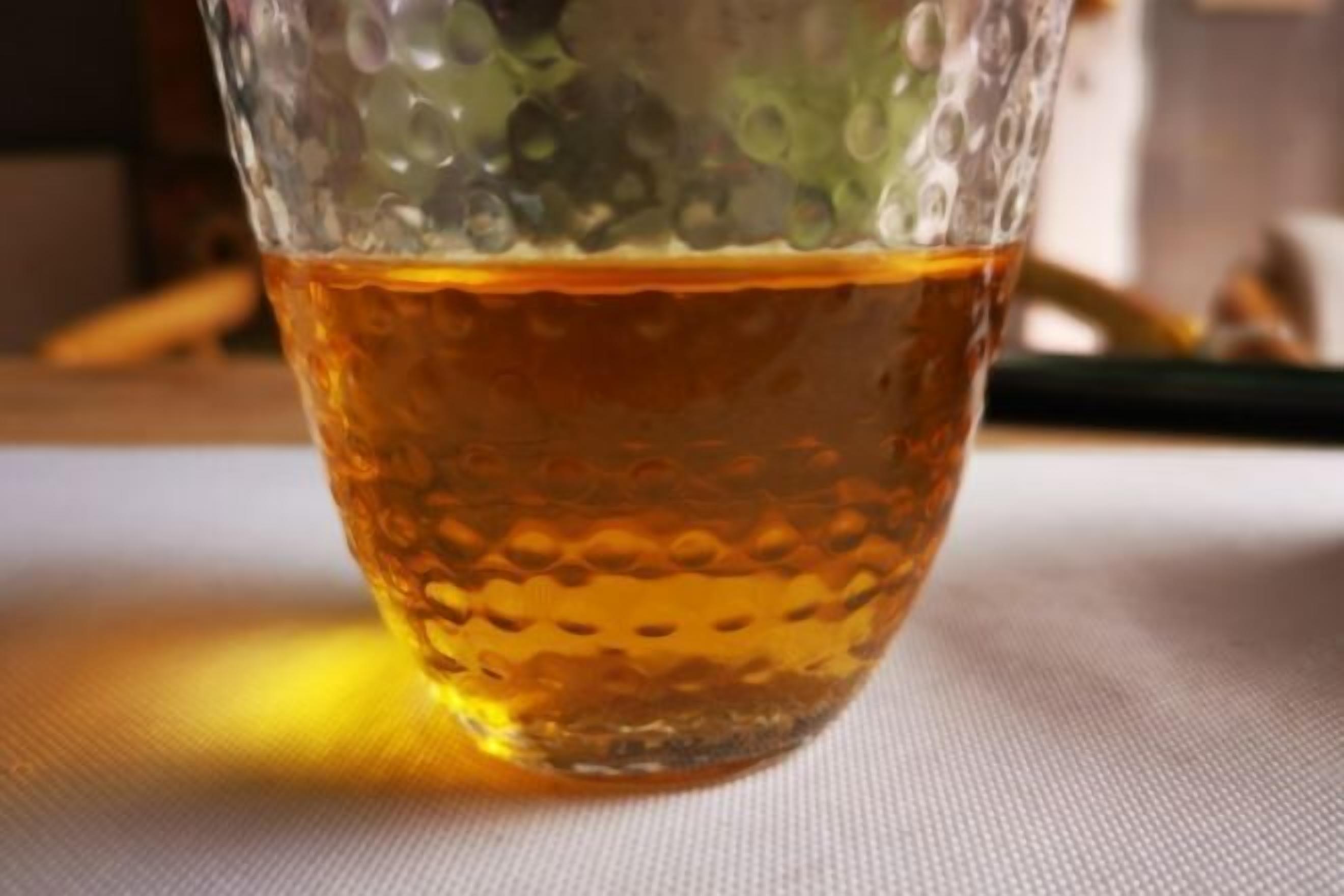 Ένα ποτήρι με ένα διαυγές, κεχριμπαρένιο υγρό, με ανάγλυφη υφή, τοποθετημένο πάνω σε μια επιφάνεια που καλύπτεται με ένα ύφασμα.