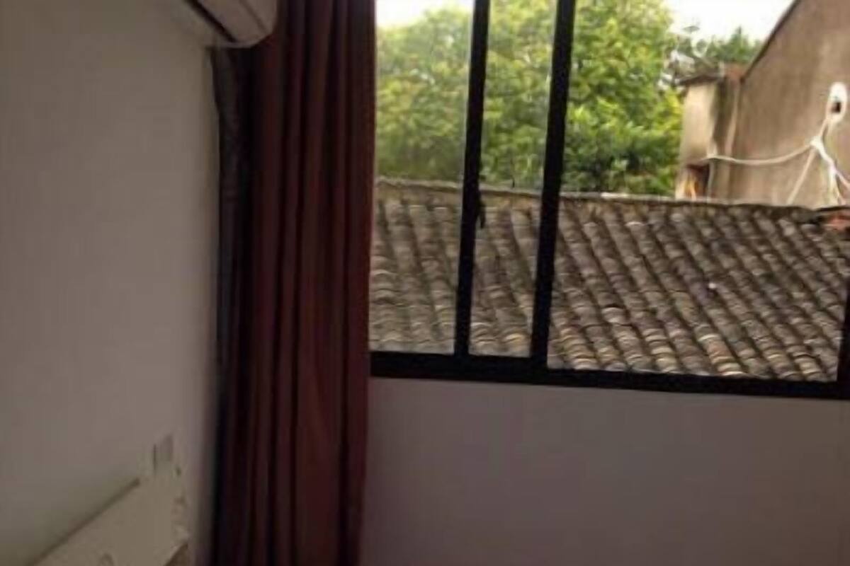 A room with a bed, a window with a view of a building with a tiled roof, and a curtain on the window.