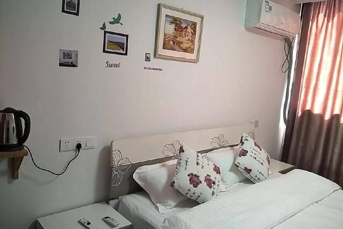 A hotel room with a bed, a bedside table, a wall-mounted air conditioner, and a window with curtains.