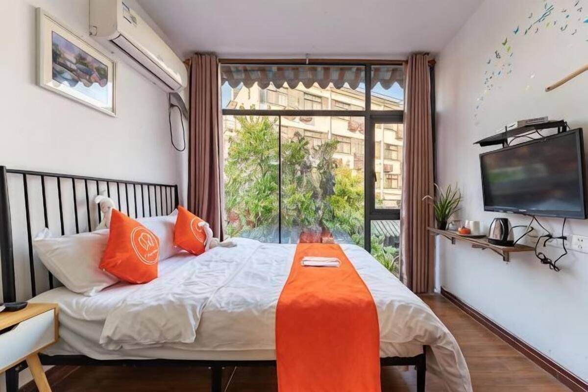 A hotel room with a bed, pillows, a blanket, a TV, and a window with a view of buildings and greenery.
