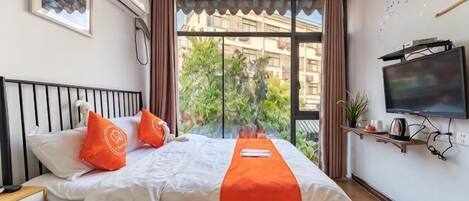 A hotel room with a bed, pillows, a blanket, a TV, and a window with a view of buildings and greenery.