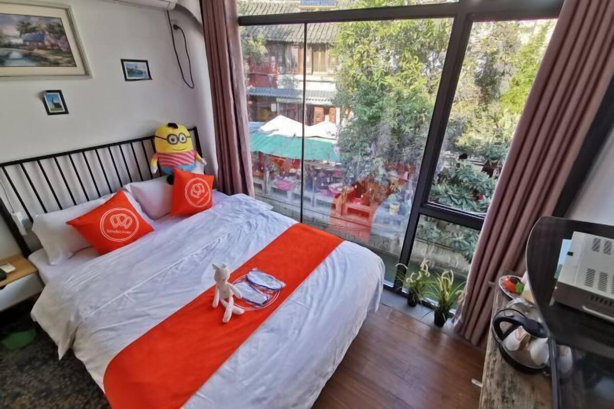 A hotel room with a large bed, a Minion plush toy, and a view of a traditional building and greenery outside.
