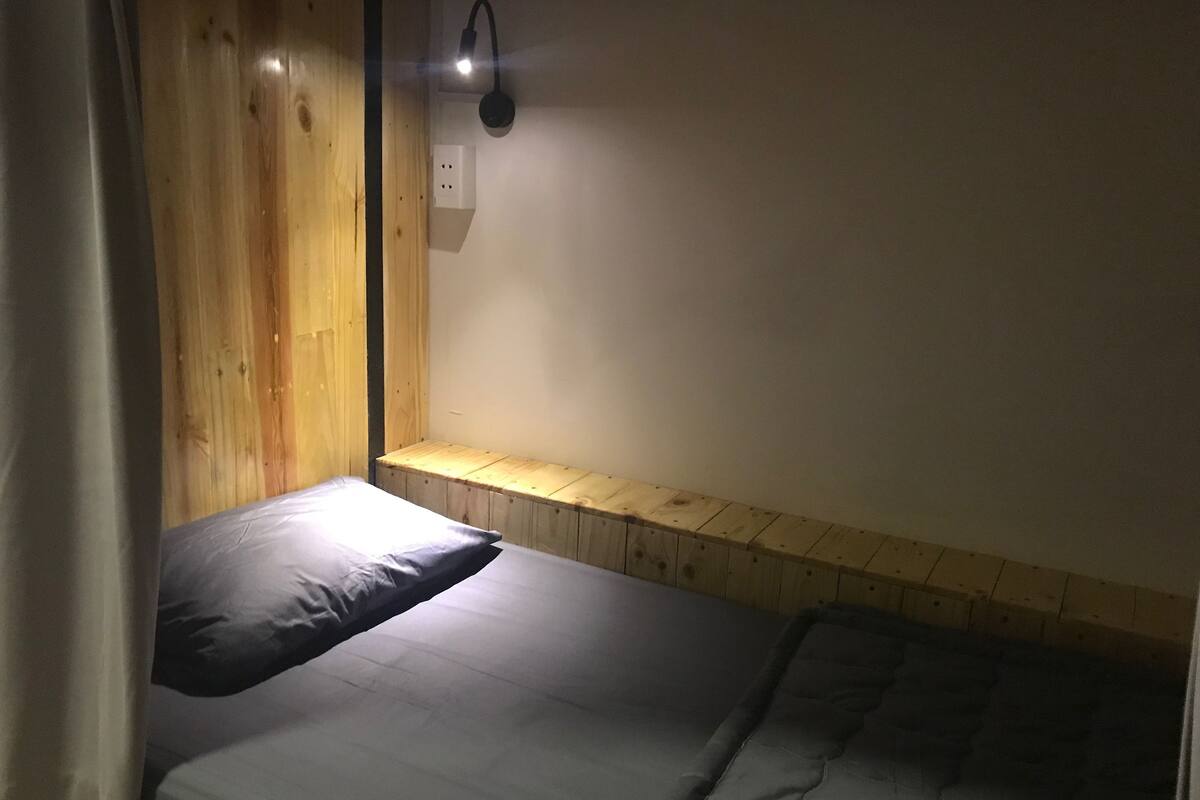 A small, single-bed room with a wooden bench and a wall-mounted light fixture.